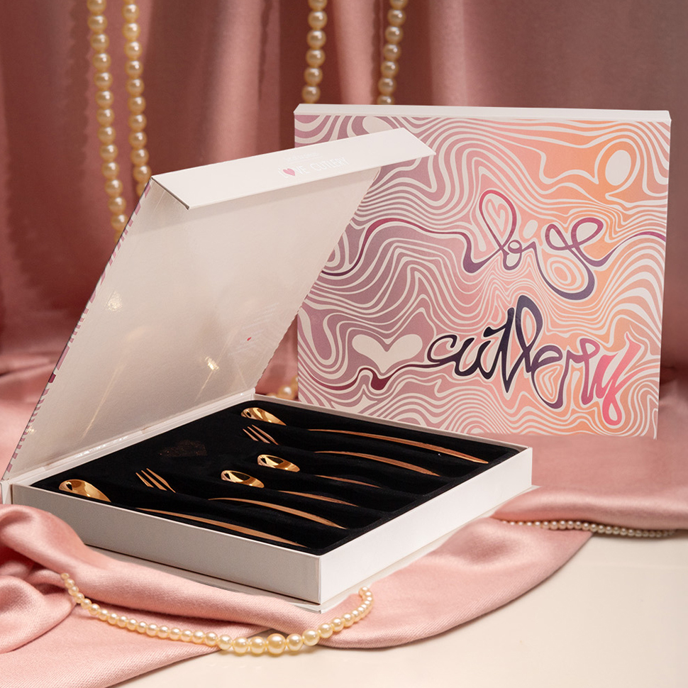 Otwarte białe pudełko ze złotymi łyżeczkami Love Cutlery położone na różowej satynie 